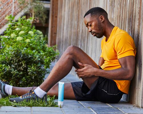 Mężczyzna w odzieży sportowej siedzący na ziemi z ugiętą nogą cierpiąc na kolano biegacza