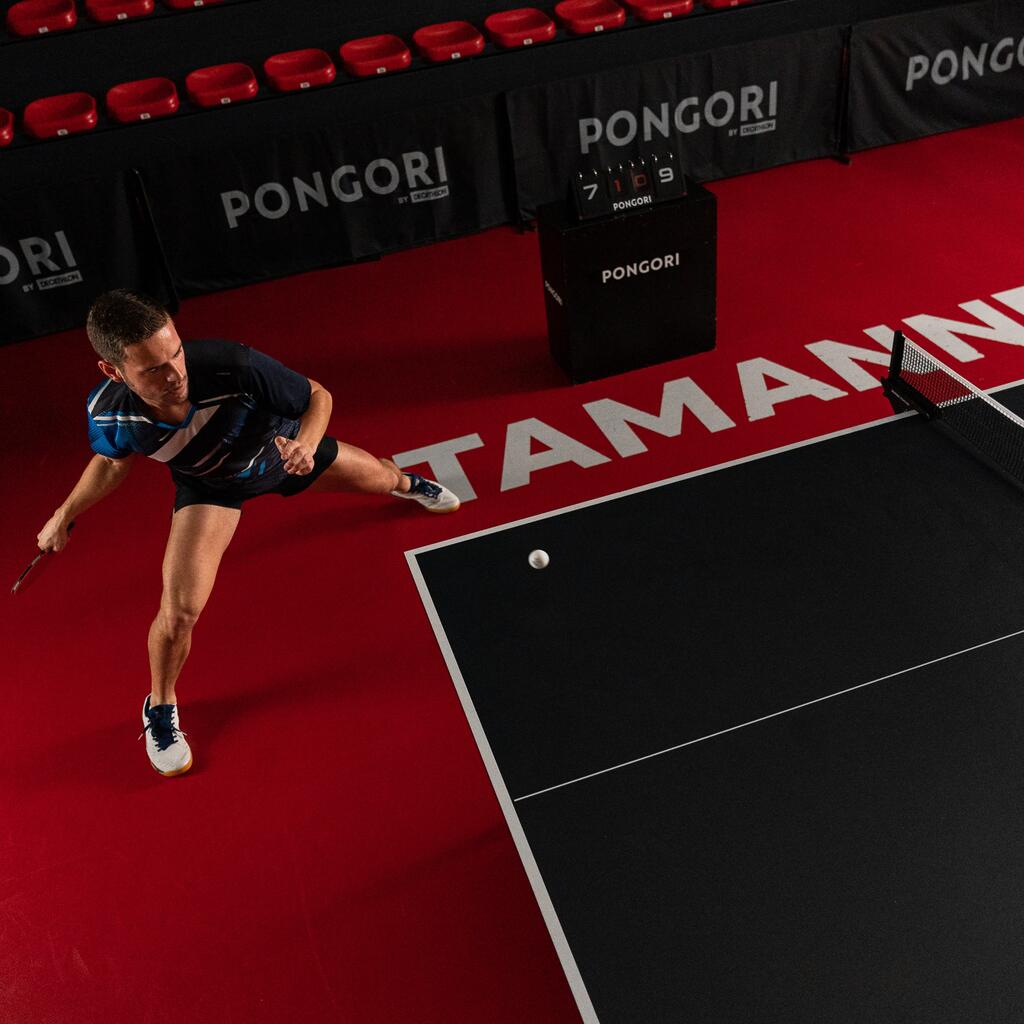 Ein Tischtennisspieler schlägt mit einem Tischtennisschläger einen Ball über einer schwarzen Tischtennisplatte in einer Indoor Halle mit rotem Boden.