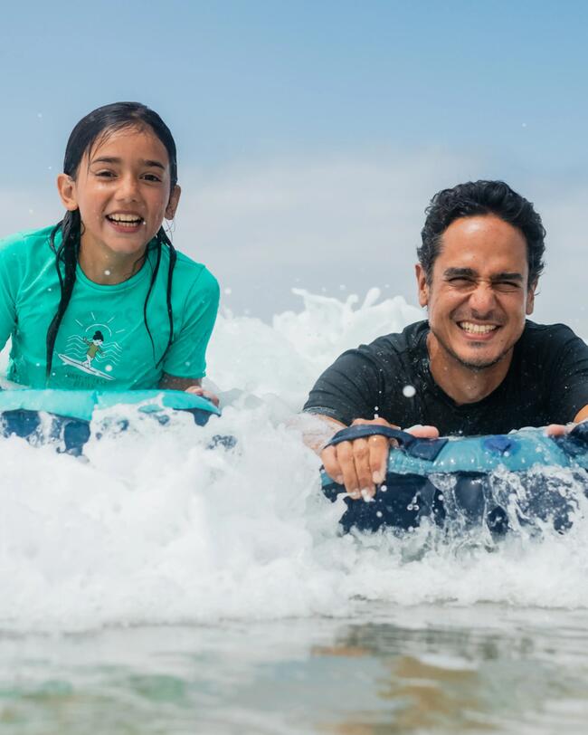 Kinder in bunten Badeanzügen laufen mit Surfbrettern am sonnigen Sandstrand entlang. Meer im Hintergrund, blauer Himmel.