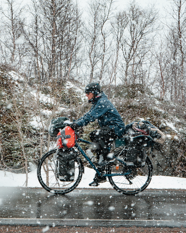Mann fährt mit blauem Reisefahrrad und Gepäck über nasse Straße; im Hintergrund Winterwald. Es schneit.