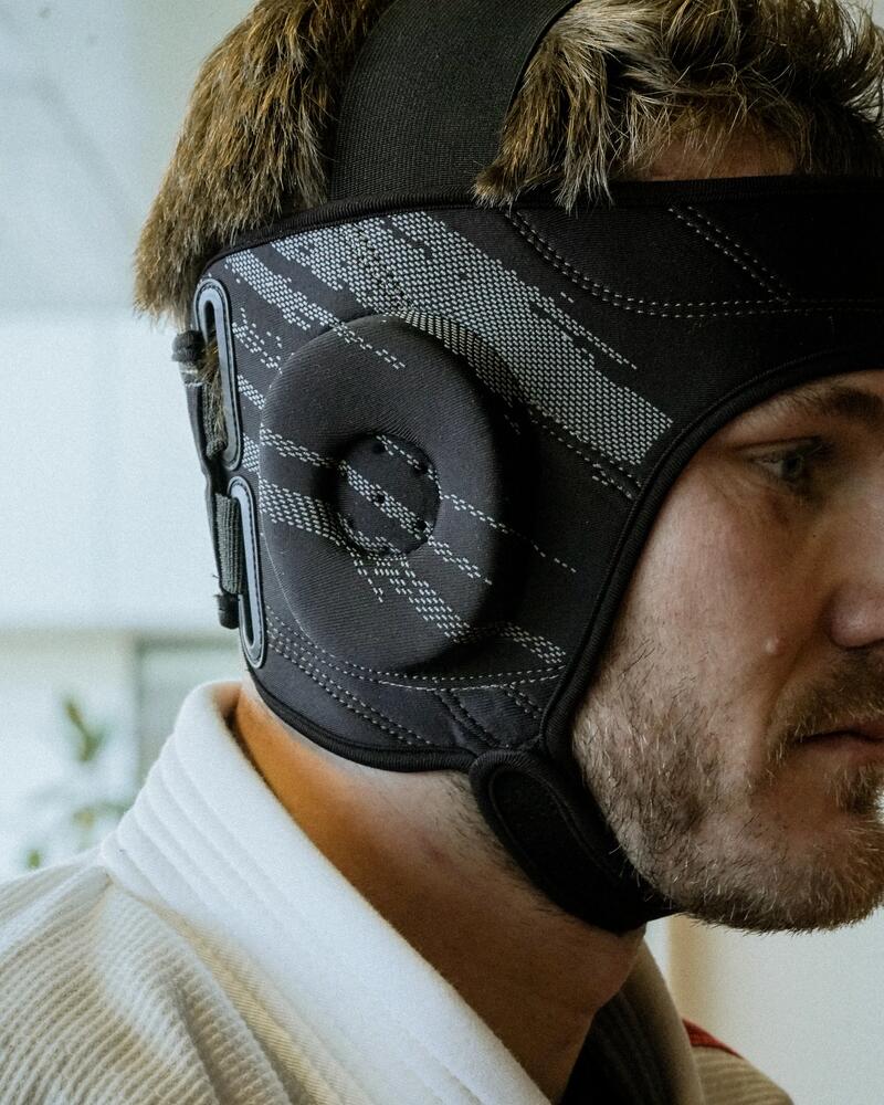 Mann mit Bart trägt einen speziellen schwarzen Kopfschutz für Kampfsport über den Ohren und einen weißen Judo-Anzug.