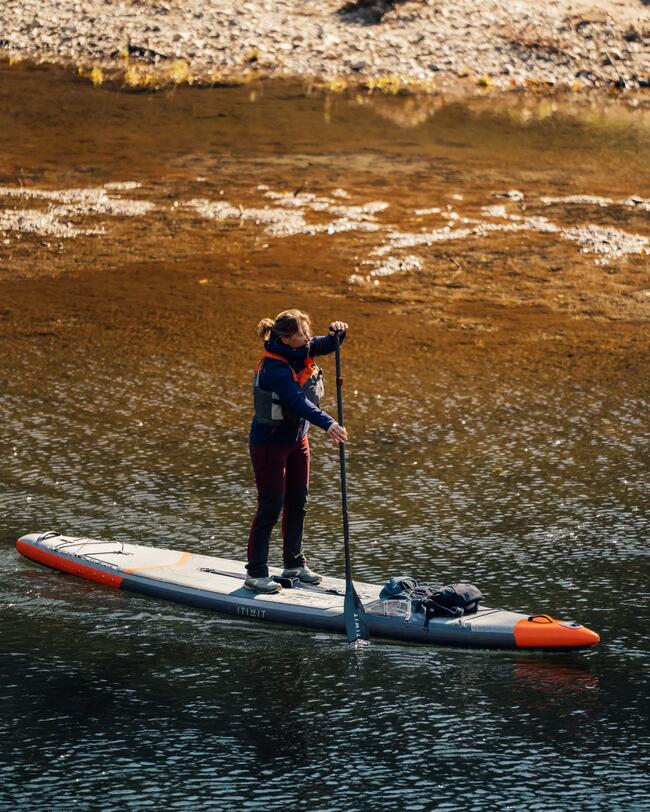 Person auf grauem Paddleboard auf ruhigem Wasser; braune und grüne Uferlandschaft im Hintergrund.