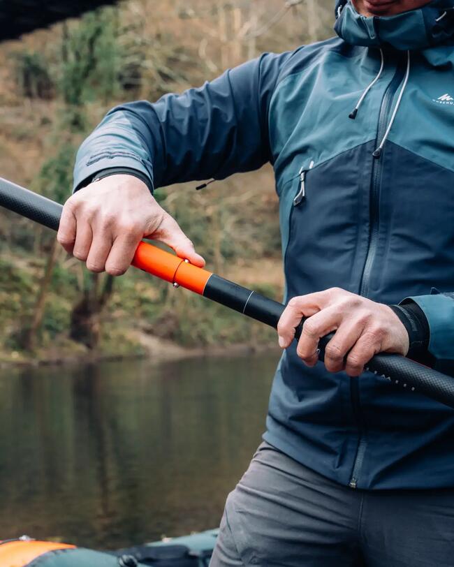 Person in blauer Jacke montiert orange-schwarzes Paddel; Hintergrund mit Wasser und unscharfer Natur.
