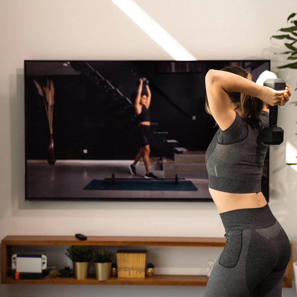 Žena vežba sa tegovima ispred televizora.