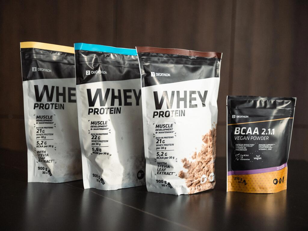 Štiri vrečke Decathlonovih prehranskih dodatkov, vključno s tremi pakiranji Whey Protein ter enim pakiranjem BCAA 2.1.1 Vegan Powder, postavljene na temni mizi.