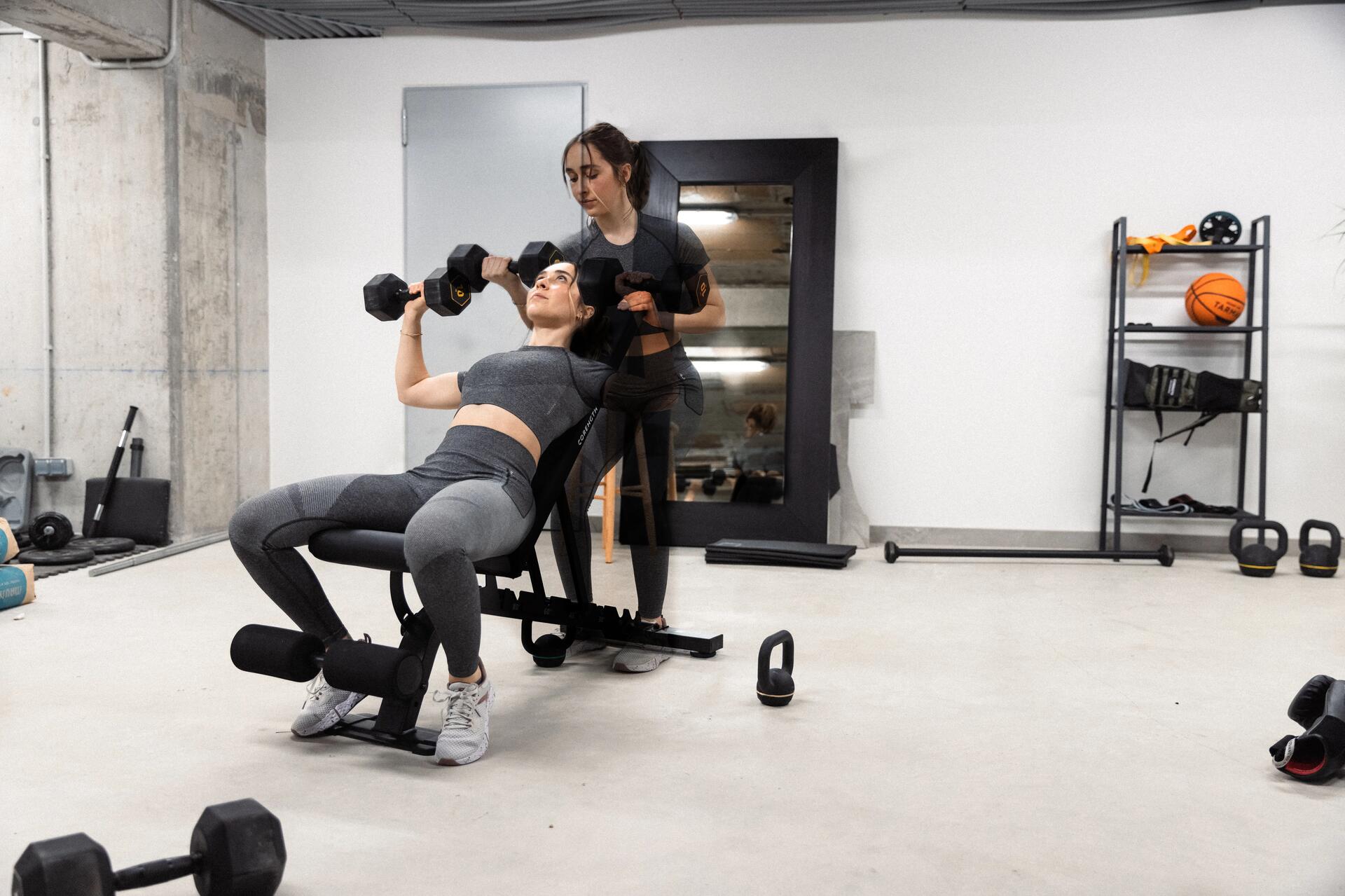 Kobieta w odzieży fitness trenująca z hantlami na ławce skośnej do ćwiczeń pod nadzorem trenerki personalnej