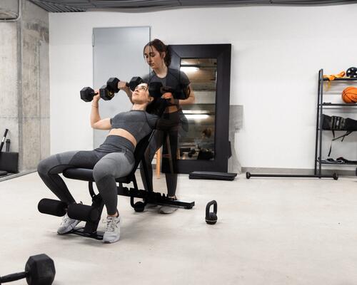 Kobieta w odzieży fitness trenująca z hantlami na ławce skośnej do ćwiczeń pod nadzorem trenerki personalnej