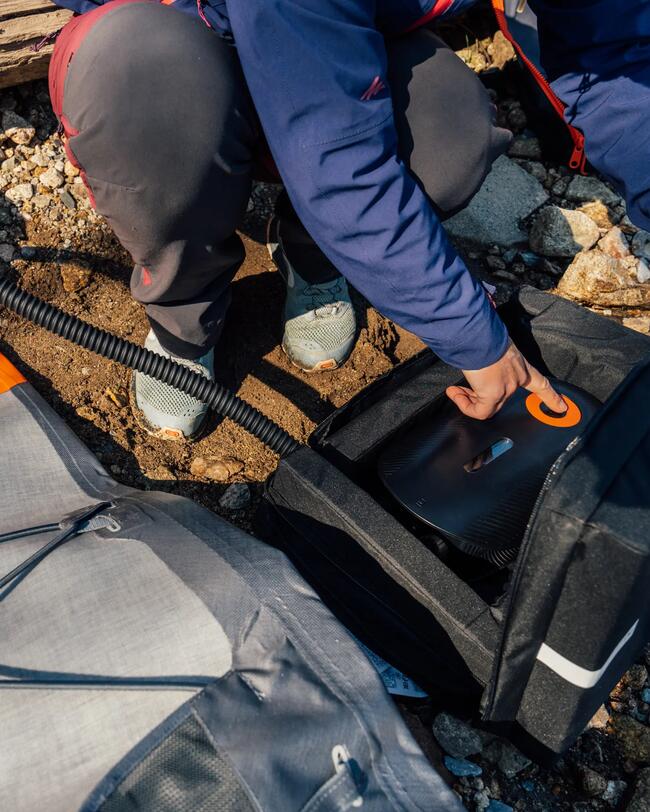 Person in blauer Jacke füllt auf steinigem Boden auf, schwarze Tasche mit orangefarbenem Detail, graue Hose, sportliche Schuhe.