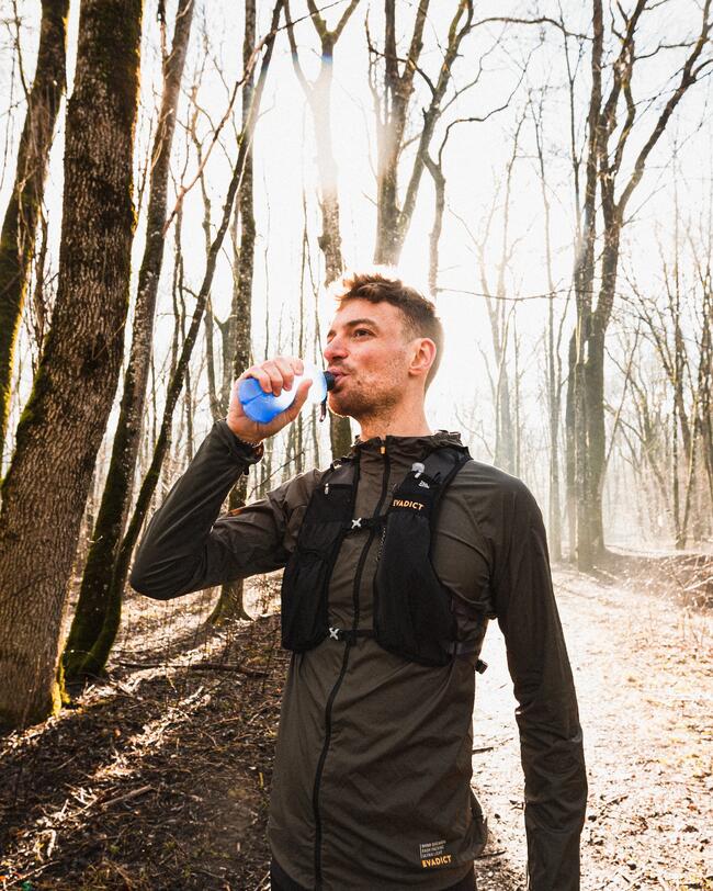 Mann in dunkler Sportkleidung trinkt aus einer Trinkflasche, umgeben von kahlen Bäumen im Sonnenlicht.