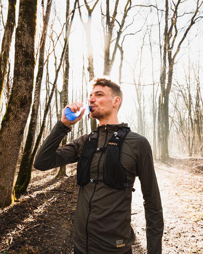 Mann trinkt aus blauer Flasche im sonnigen Wald, Bäume werfen lange Schatten, brauner Boden, klare Herbstluft.