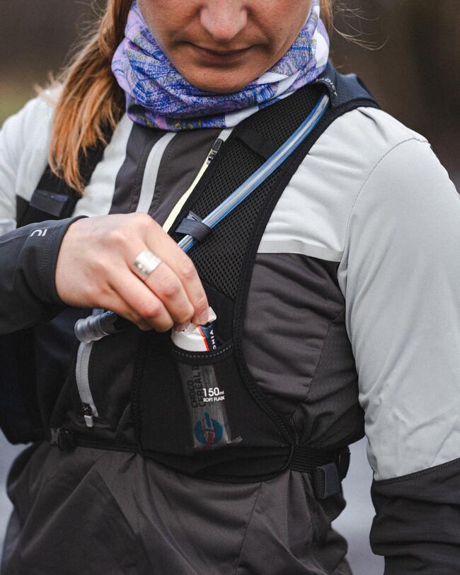 Frau steckt Riegel in schwarze Weste, graue Jacke, blauer Schal, Outdoor-Ausrüstung, energisch, sportlich.