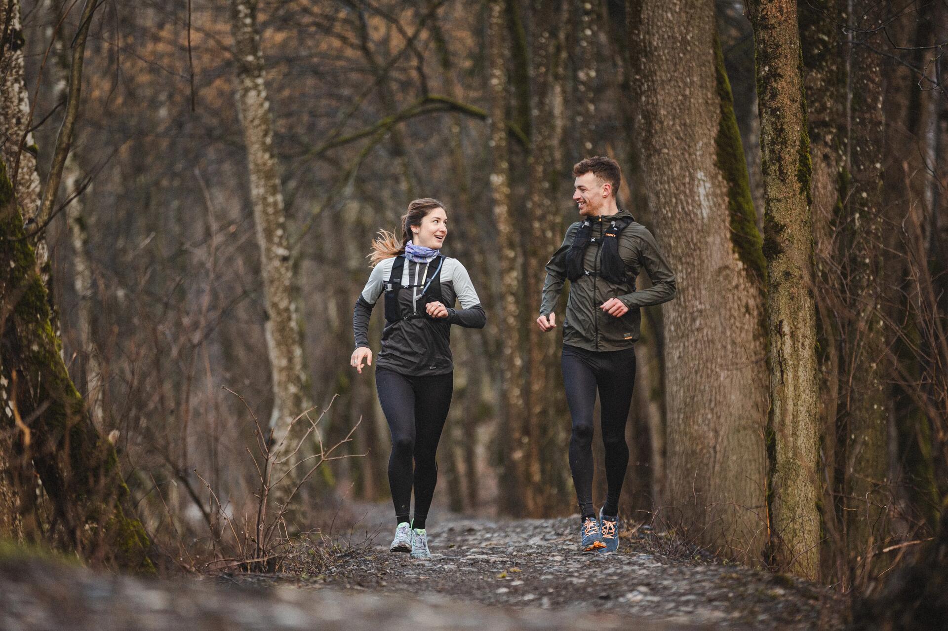 Zwei junge Menschen joggen auf grauem Waldweg, umgeben von braunen Bäumen und naturbelassenen Strukturen.