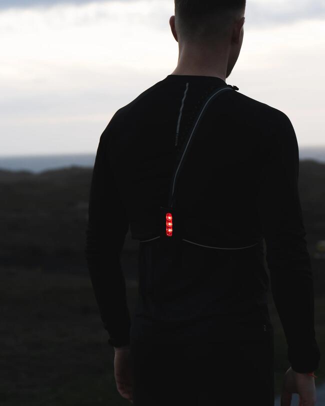 Person in dunkler Kleidung mit roten LEDs auf dem Rücken, steht in ruhiger Landschaft bei Dämmerung.