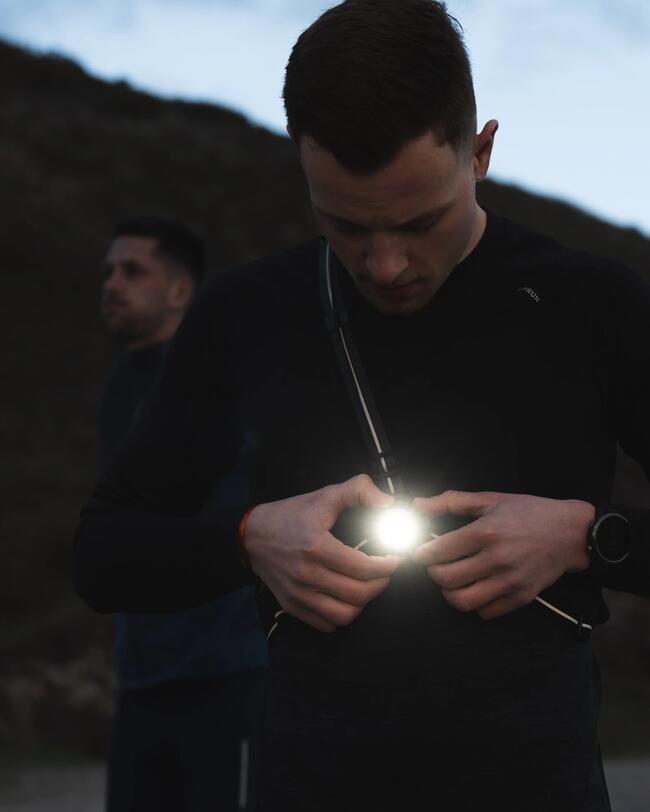 Mann im Dunkeln mit leuchtender Stirnlampe, dunkle Kleidung, unscharfer Hintergrund mit Naturkulisse.