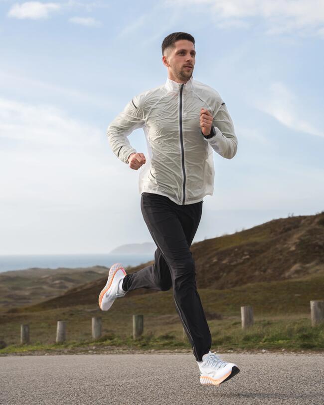Mann joggt in weißer Jacke und schwarzen Hosen auf einer Straße, blauer Himmel mit Wolken im Hintergrund.