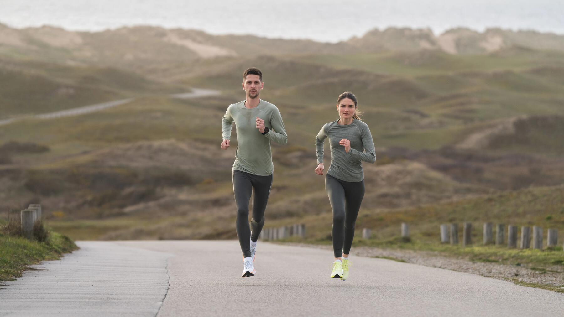 Zwei Menschen joggen auf einer Straße vor grüner Hügellandschaft, tragen sportliche Kleidung in Grau und Grün.