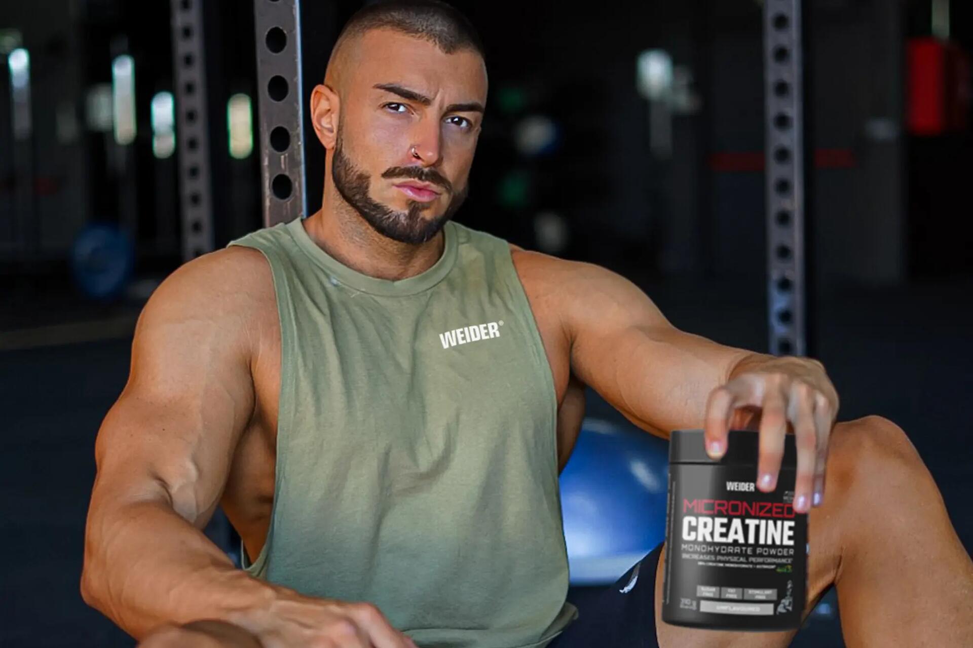 Mężczyzna w odzieży fitness trzymający w dłoni opakowanie z kreatyną siedząc na podłodze w siłowni