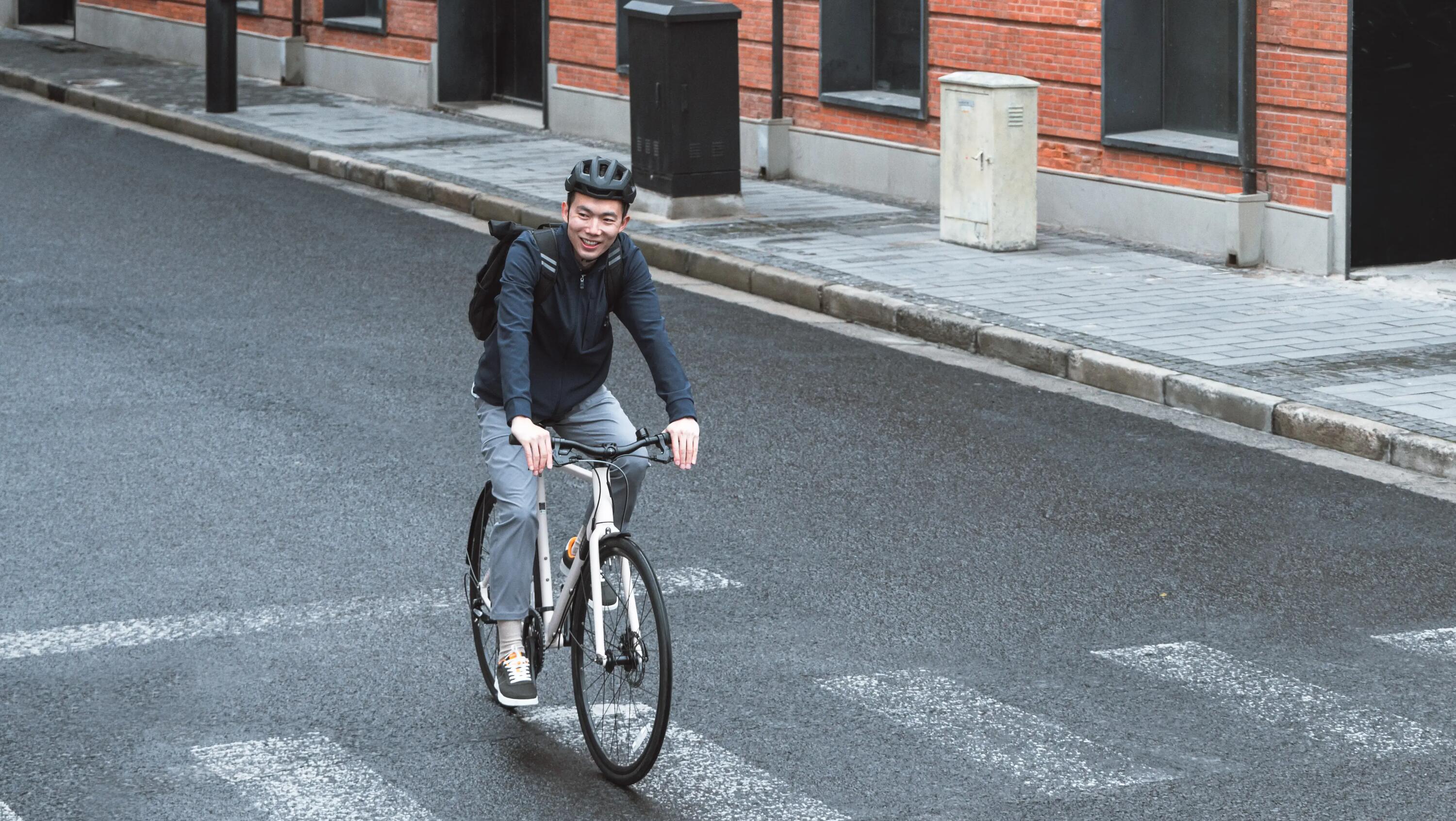 Ein urbaner Pendler mit Helm, dunkler Jacke und Rucksack überquert auf einem weißen, minimalistischen City Speed Bike (Modell Speed 500) einen Zebrastreifen. Die Szene spielt in einer ruhigen Stadtstraße vor einer markanten, roten Backsteinfassade mit großen Fensteröffnungen bei Tageslicht.
