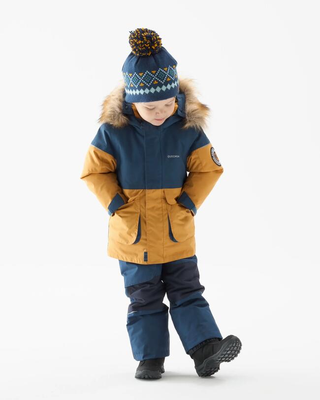 Ein Junge in orange-blauer Winterjacke, blauer Winterhose, schwarzen Winterschuhen und blau gemusterter Bommelmütze steht mit den Händen in der Jackentasche vor einer grauen Leinwand und schaut auf seinen linken Fuß, den er gerade anzieht.