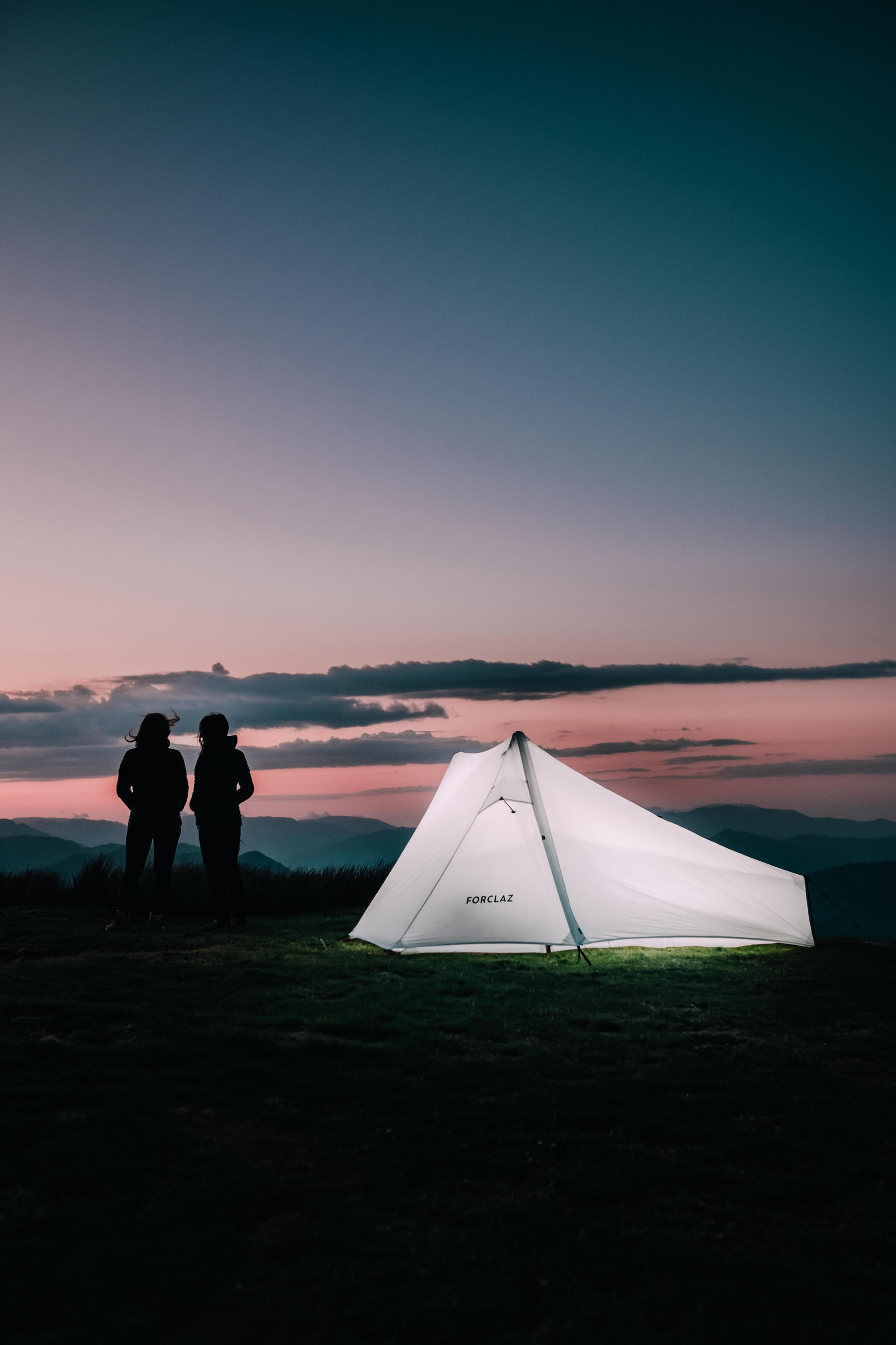 Dwie osoby podziwiające zachód słońca stojące obok rozbitego namiotu dwuosobowego