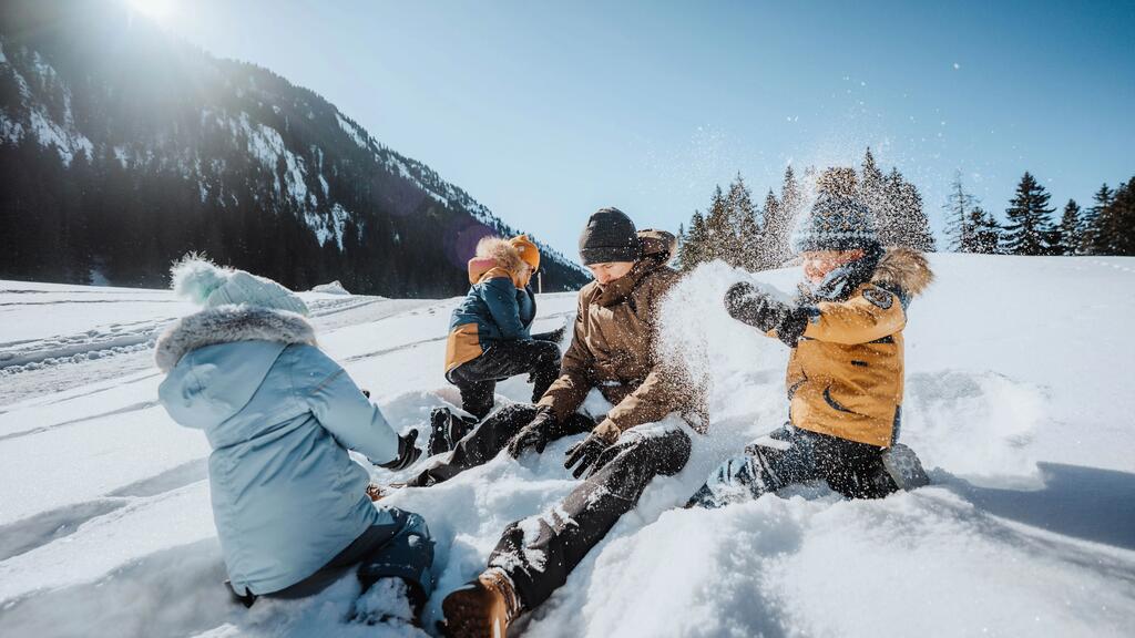 Vier Kinder in bunten Winterjacken spielen lachend im weißen glitzernden Schnee unter klarem, sonnigem Himmel und vor Bergen.