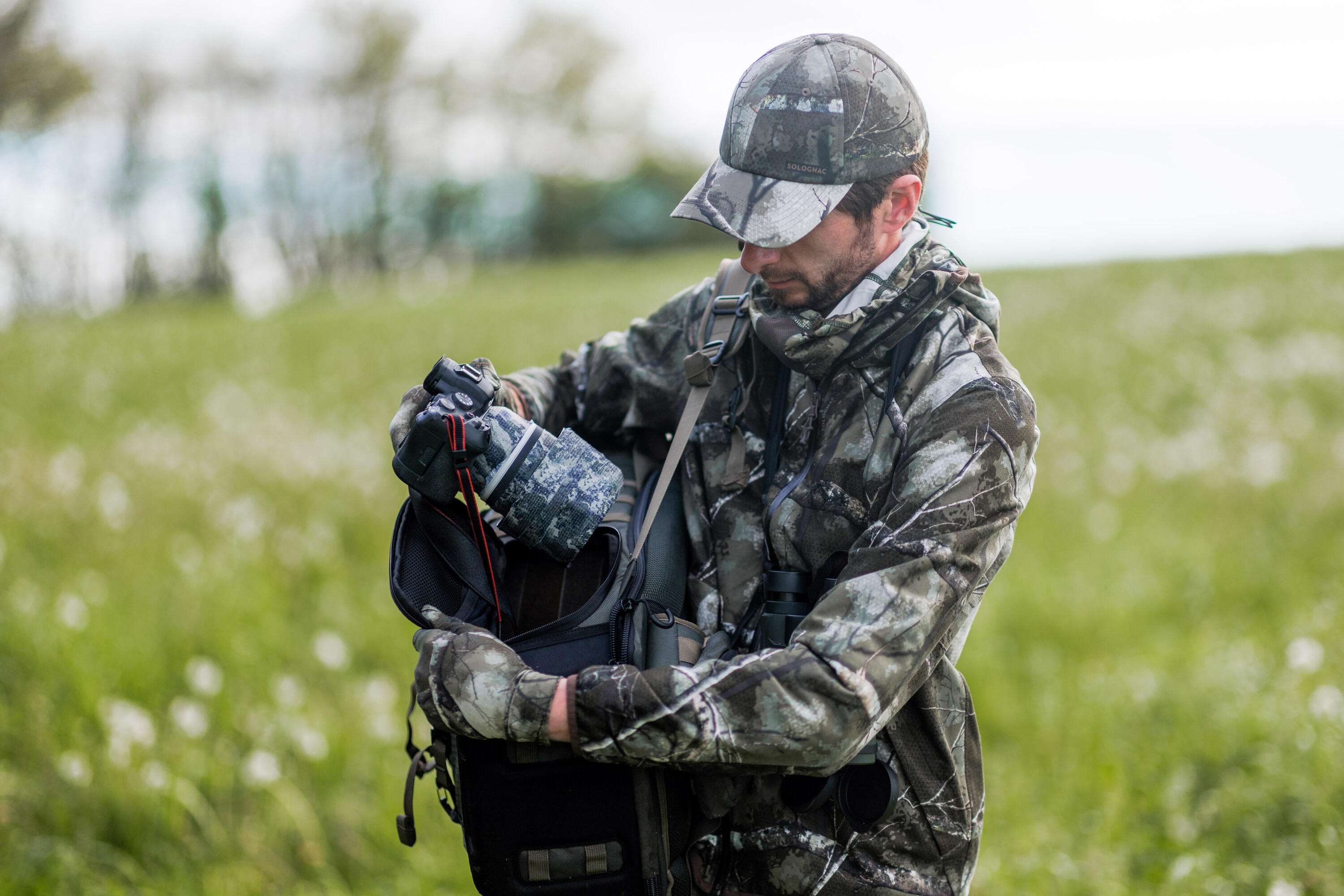 un photographe animalier avec une tenue Treemetic sort son appareil de son sac au milieu d'une prairie