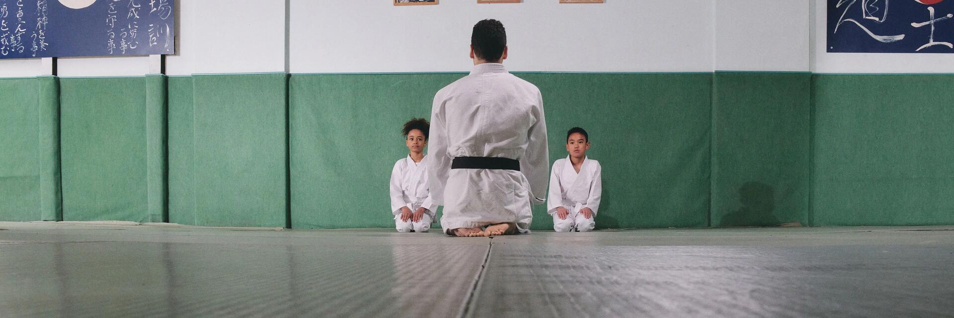 Dzieci siedzące na podłodze w strojach do karate z trenerem opowiadającym o zasadach sztuk walki