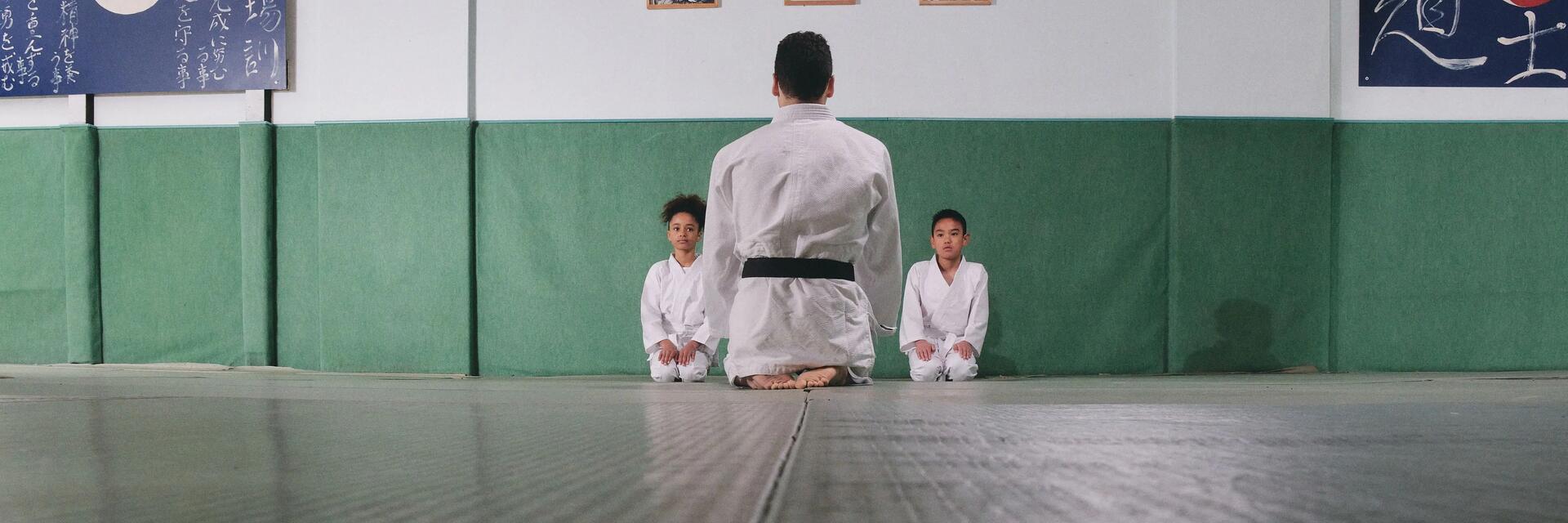 Dzieci siedzące na podłodze w strojach do karate z trenerem opowiadającym o zasadach sztuk walki 