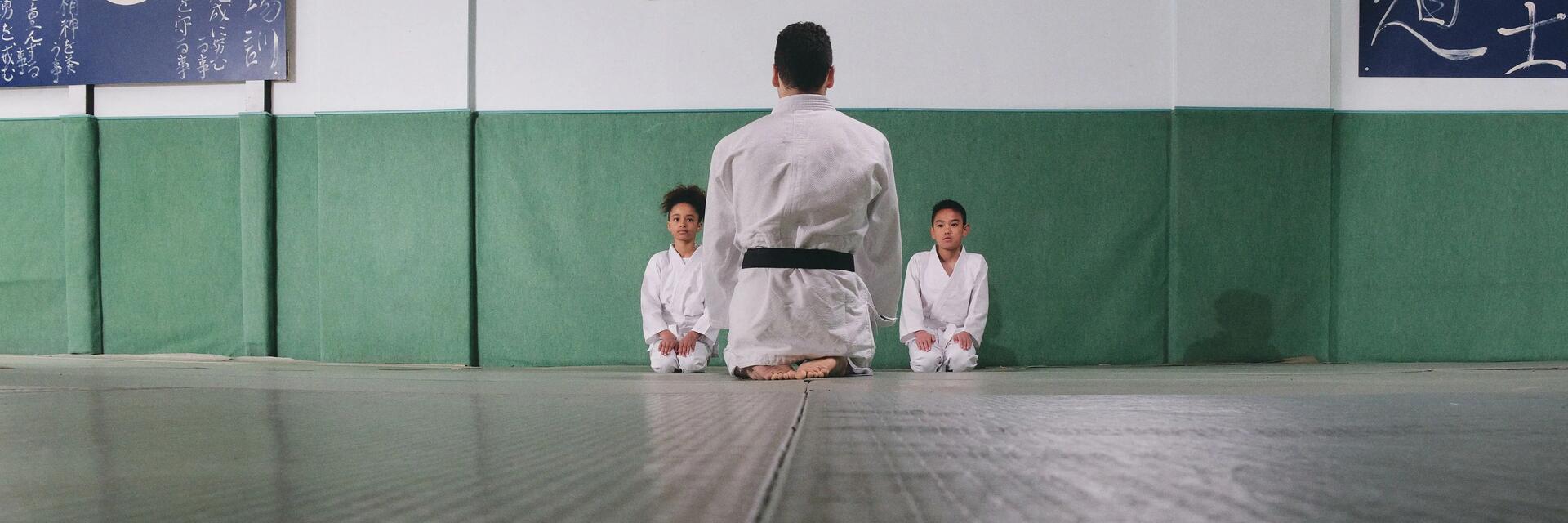 Dzieci siedzące przy ścianie przed nimi trener judo