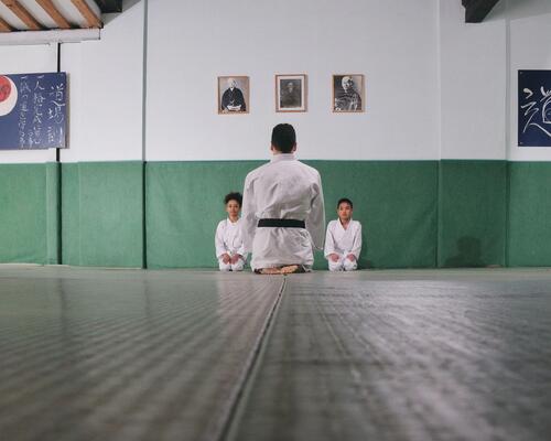 Dzieci siedzące na podłodze w strojach do karate z trenerem opowiadającym o zasadach sztuk walki 