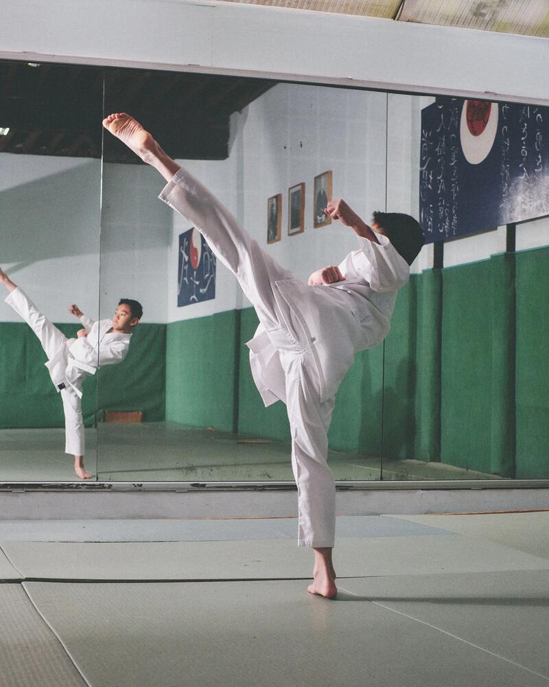 Junge im Karateanzug führt einen hohen Tritt vor einem Spiegel in einem Dojo aus.