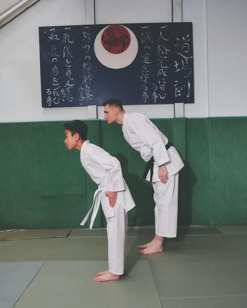 Ein Schüler und ein Lehrer in Karateanzügen verbeugen sich gemeinsam in einem Dojo vor einer Tafel mit Schriftzeichen.