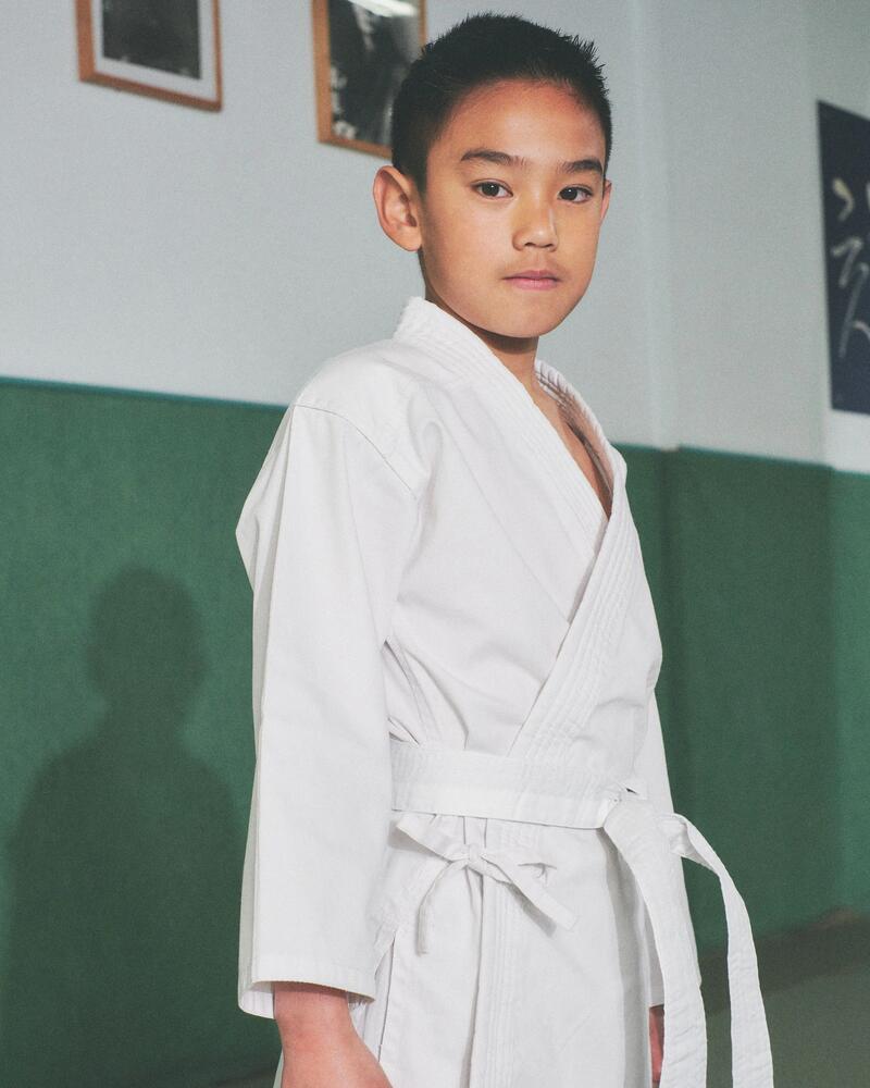 Ein Junge in einem weißen Kampfsportanzug mit weißem Gürtel steht in einem Dojo vor einer grünen Wand.