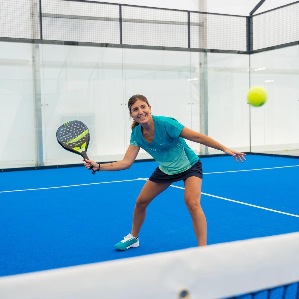  Eine lachende Padel-Spielerin in einem blau-grünen Oberteil und einem schwarzen Rock schlägt einen Ball auf einem blauen Padel-Platz.