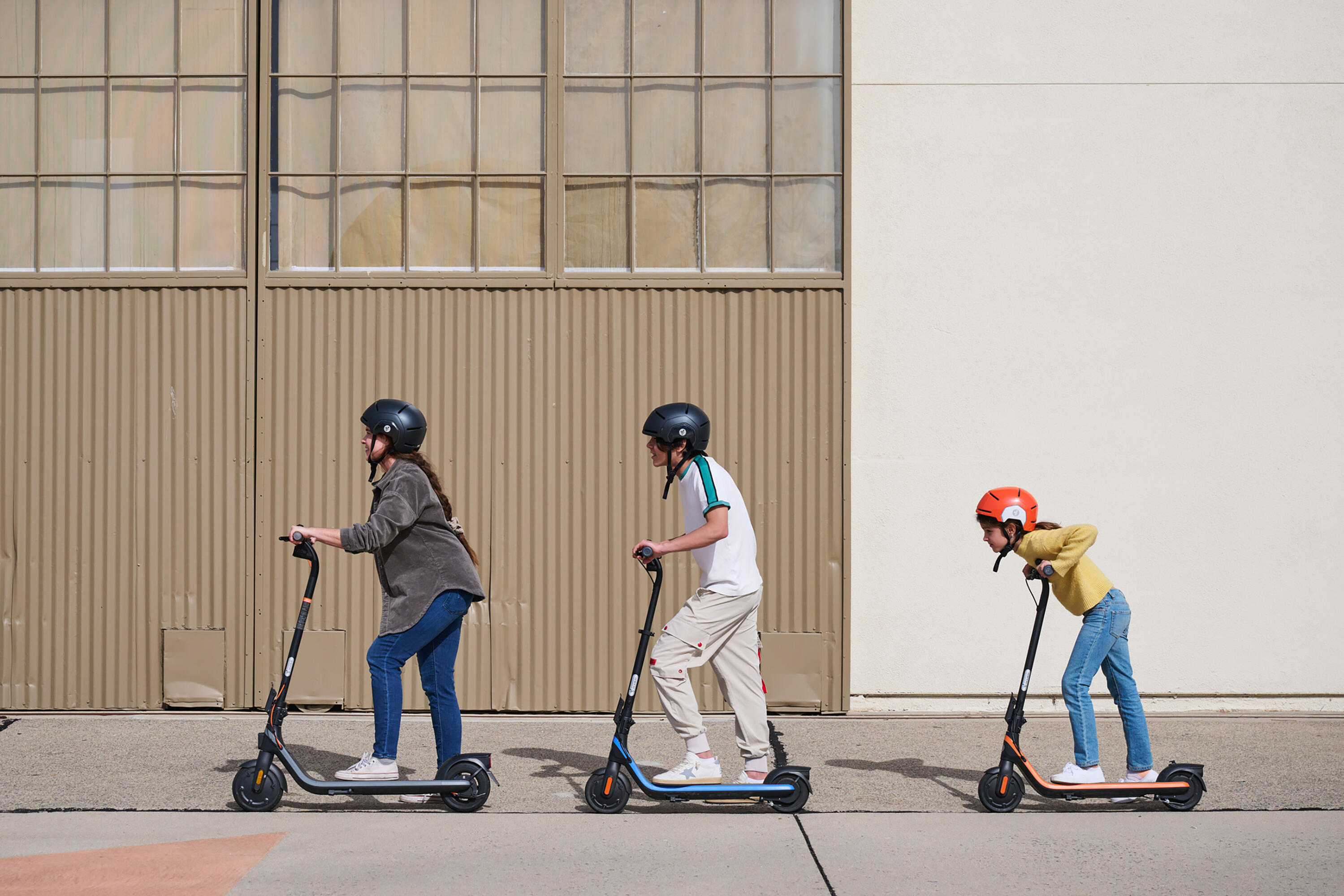 Three helmeted riders travelling on electric scooters along an urban street.