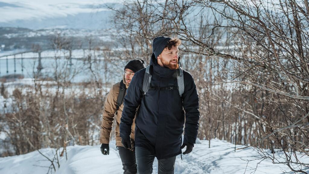 Zwei Männer in dunkler Winterkleidung wandern durch verschneite Landschaft mit kahlen Bäumen und Bergen im Hintergrund.