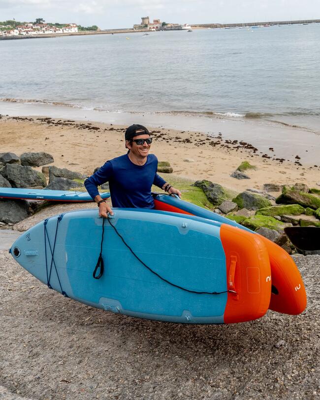 Mann in Blau mit Paddleboards, bunt und rund, Sandstrand, graue Felsen, Meer im Hintergrund, wolkiger Himmel, entspannte Stimmung.
