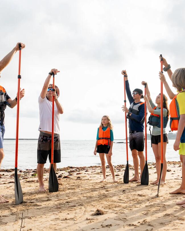Gruppe Menschen mit Schwimmwesten hält Paddel am Sandstrand; bedeckter Himmel, ruhiges Meer.