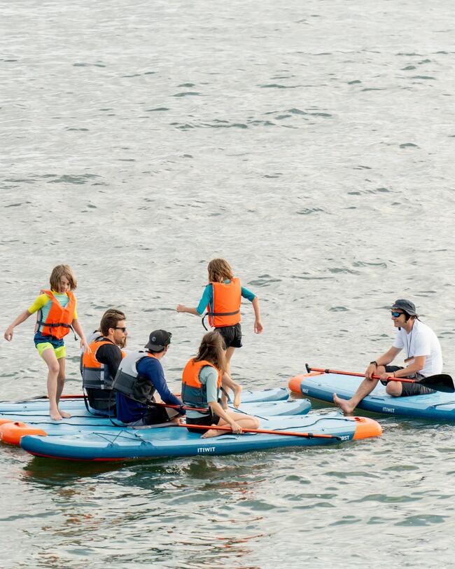 Mehrere Personen mit Schwimmwesten auf blauen Stand-Up-Paddle-Boards auf ruhigem Wasser. Kinder spielen und Erwachsene sitzen.
