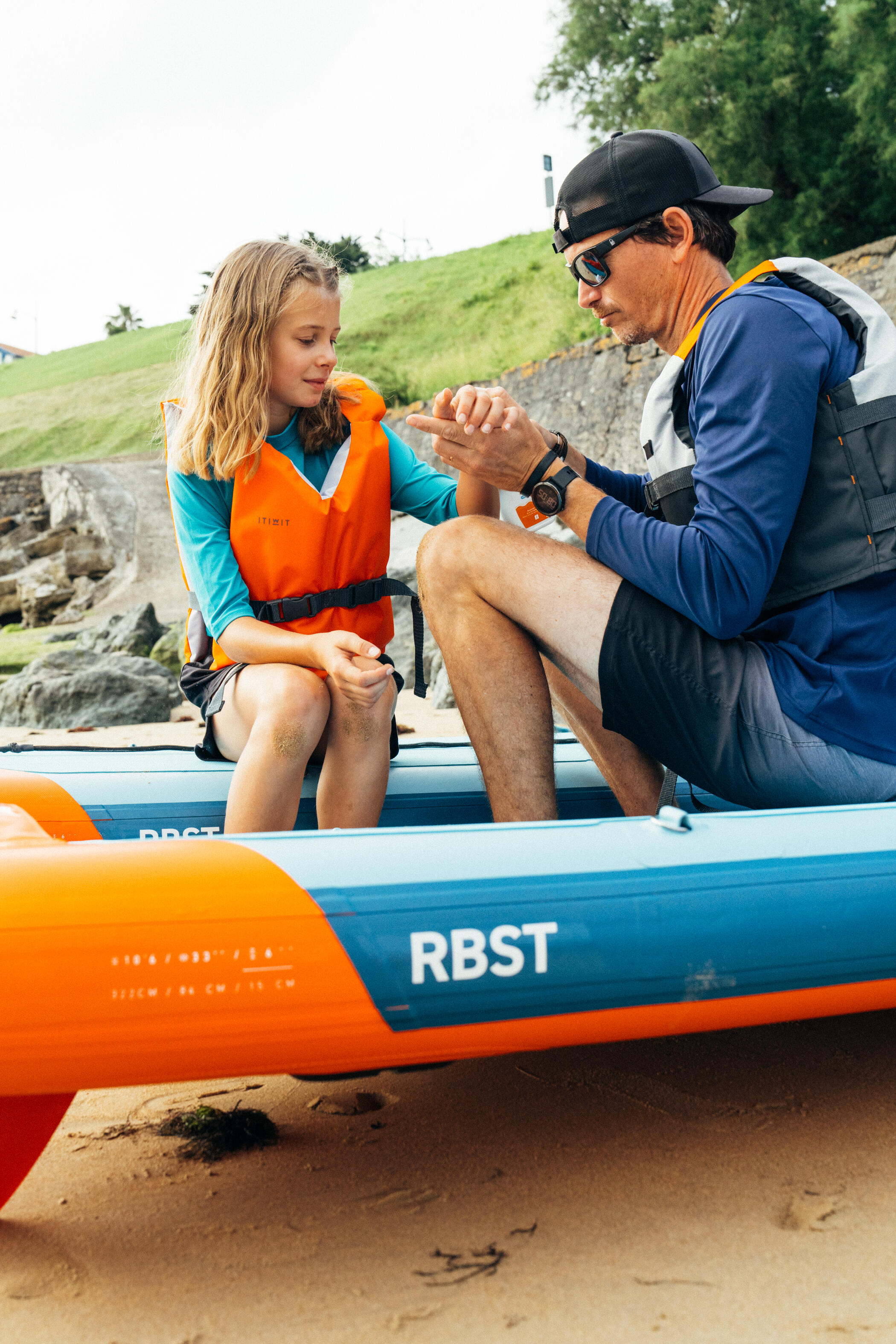 Mann und Kind sitzen an Land auf einem SUP mit Schwimmwesten