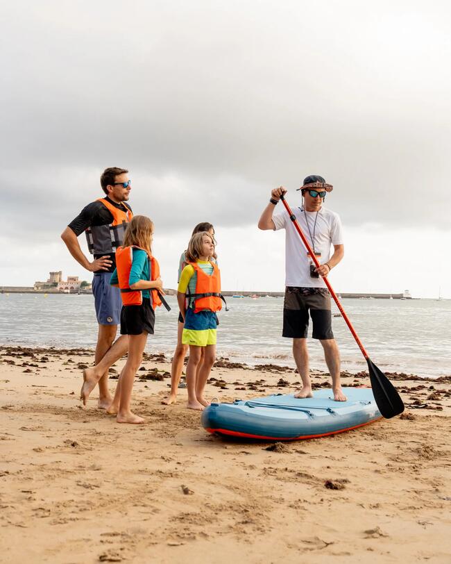 Kinder stehen am Strand vor einem blauen SUP, während ein Mann das Paddeln demonstriert. Ruhiges Wasser und bewölkter Himmel mit Küste.