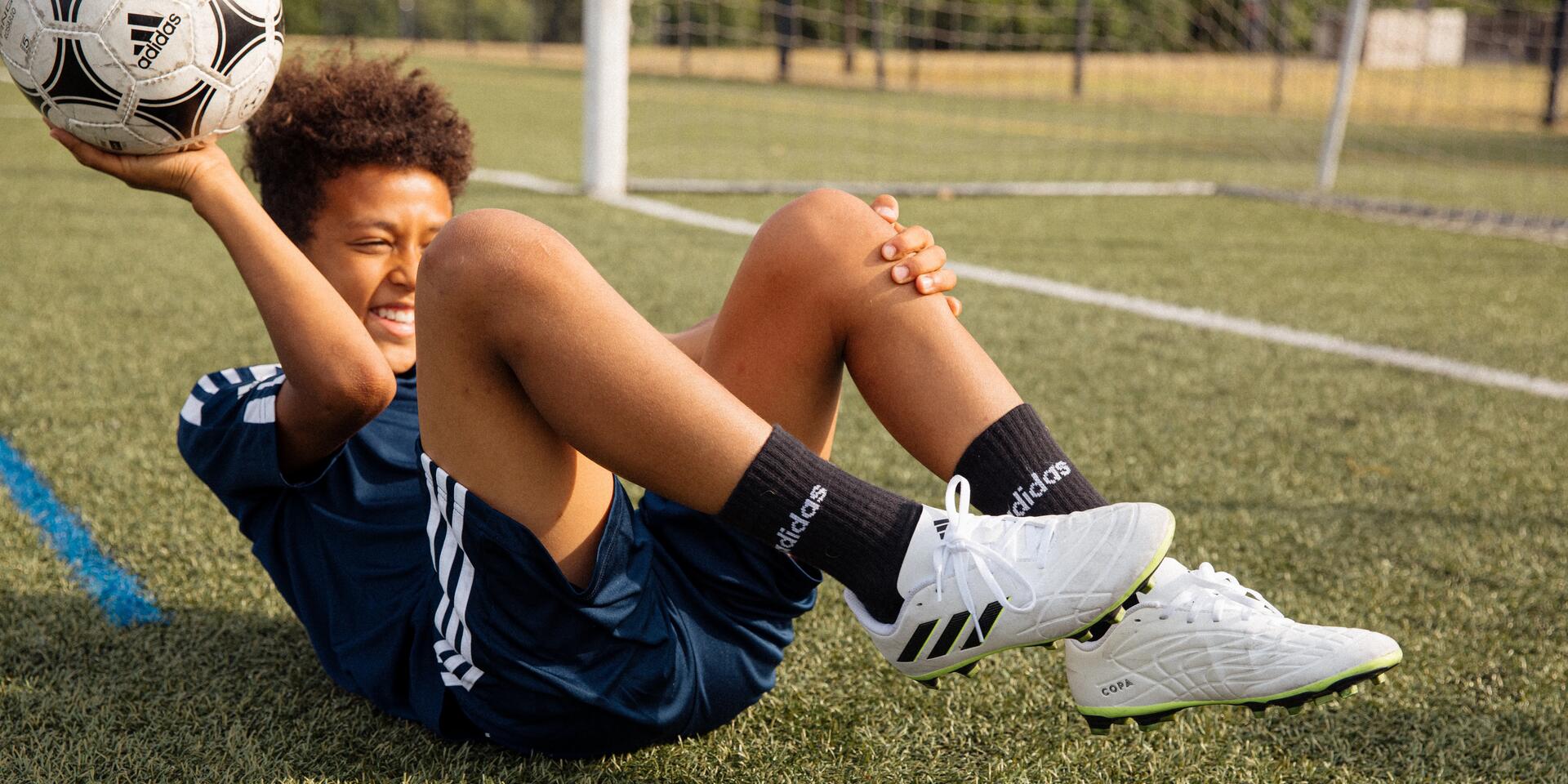 chłopiec w butach piłkarskich trzymający piłkę do gry w piłkę nożną siedząc na murawie