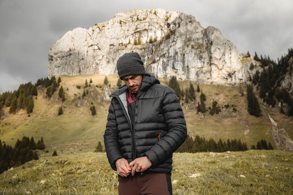 Moški s temno sivo kapo zapenja črno puhasto jakno s kapuco na gorskem travniku pred veliko skalnato goro.