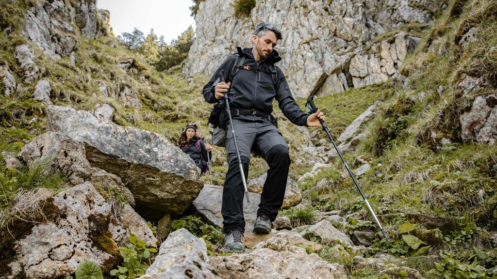Zwei Wanderer in dunkler Outdoor-Kleidung gehen mit Stöcken durch grünes, felsiges Gebirge mit steilen, grauen Felshängen.