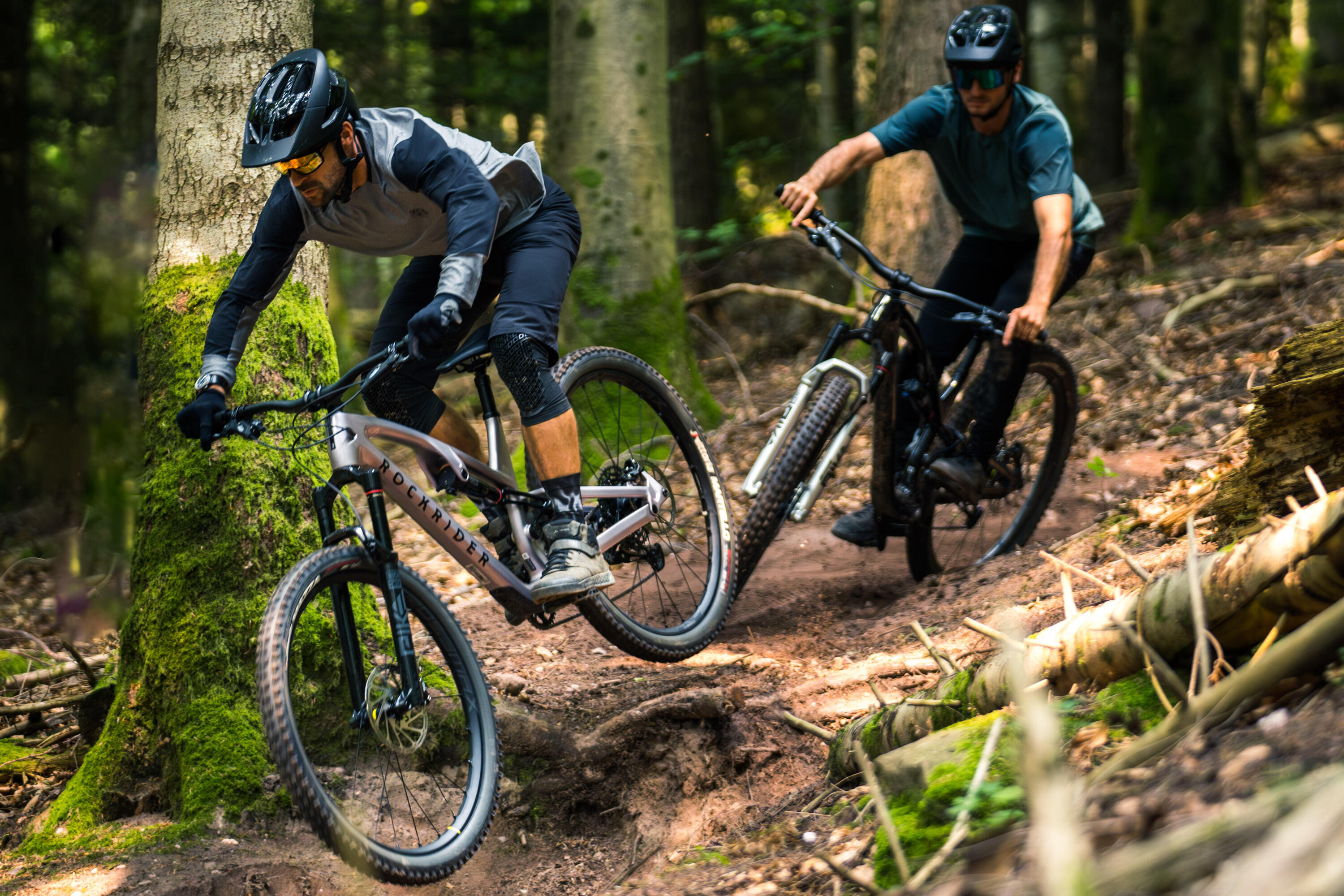 Zwei Mountainbiker mit Helmen fahren hintereinander einen unebenen Pfad durch einen dichten Wald hinab. Der vordere Fahrer trägt ein grau-schwarzes Oberteil, während der hintere ein blaugrünes T-Shirt trägt.