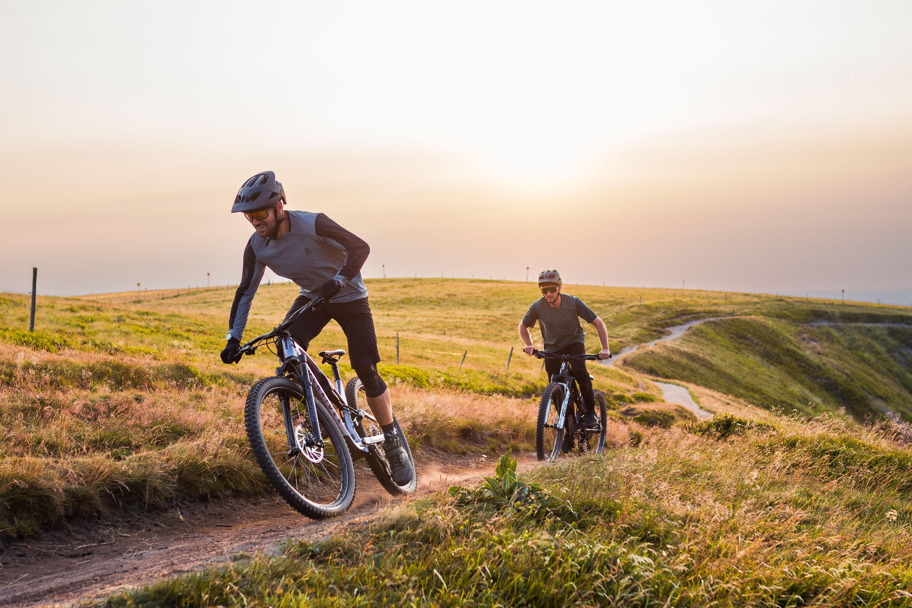 Zwei Mountainbiker fahren bei warmem Licht des Sonnenuntergangs auf einem Pfad über eine hügelige, von Gras bewachsene Wiese.