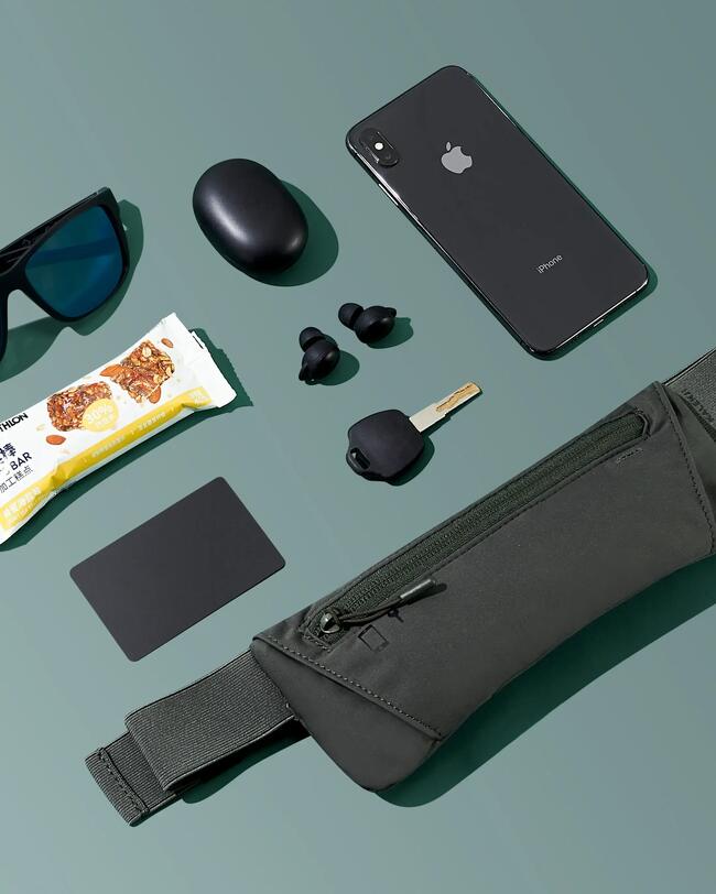 Gürteltasche, schwarze Sonnenbrille, Handy, Riegel, Ohrstöpsel und Schlüssel auf einem grünen Hintergrund.