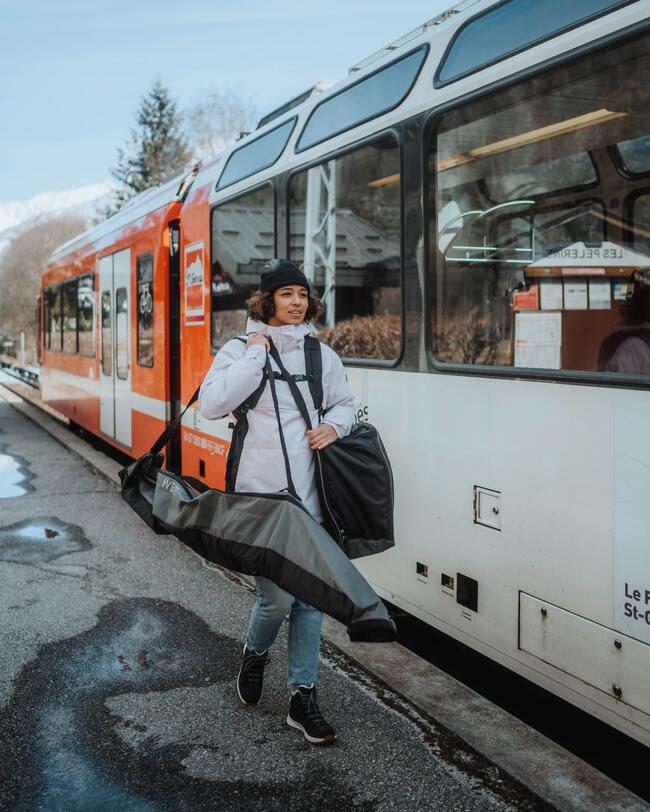 Eine Frau mit Rucksack, Skitasche und Skischuhtasche über ihren Schultern läuft neben einem stehenden Zug entlang.