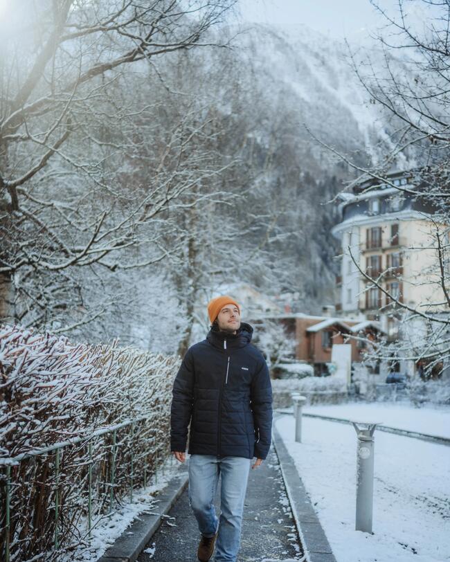Ein Mann trägt die Ski- und Snowboardjacke Herren 100 blau mit einer orangenen Mütze und läuft dabei über einen Fußgängerweg.