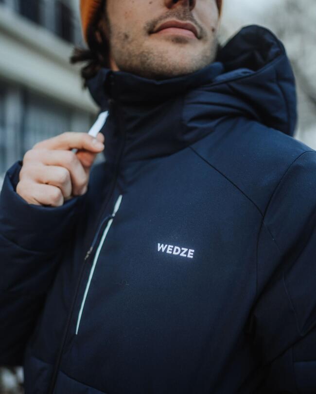 Ein Mann trägt die Ski- und Snowboardjacke Herren 100 blau, wobei der Fokus auf den reflektierendem Logo und dem reflektierenden Reißverschluss liegt.